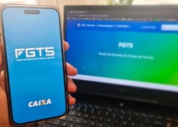 Mão segurando celular com tela do FGTS e notebook ao fundo exibindo site da CAIXA