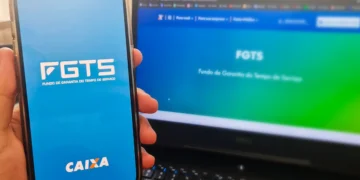 Mão segurando celular com tela do FGTS e notebook ao fundo exibindo site da CAIXA