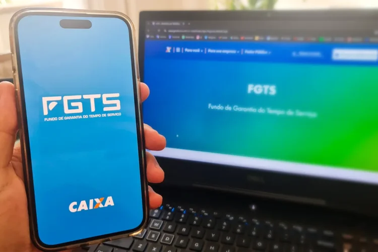 Mão segurando celular com tela do FGTS e notebook ao fundo exibindo site da CAIXA
