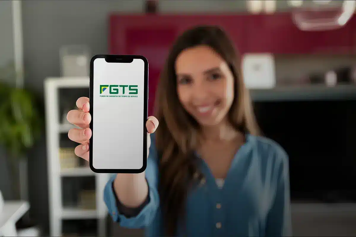 Saque de R$ 6.220 da CAIXA é liberado para cidades de São Paulo e do Rio de Janeiro a partir desta quarta-feira (18/03) 2 Mulher sorridente segurando smartphone exibindo o logotipo oficial do FGTS em uma sala.