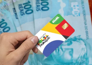 Mão segurando cartão do Programa Bolsa Família na frente de notas de cem reais representando o abono natalino proposto para 2026