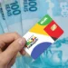 Projeto que prevê 13º do Bolsa Família volta a gerar expectativa para 2026 9 Mão segurando cartão do Programa Bolsa Família na frente de notas de cem reais representando o abono natalino proposto para 2026