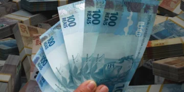 Mão segurando notas de R$ 100 com vários maços de dinheiro ao fundo