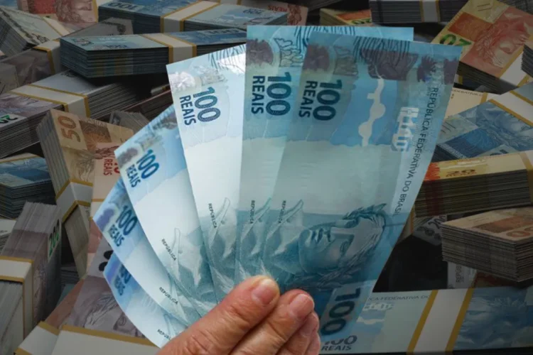 Mão segurando notas de R$ 100 com vários maços de dinheiro ao fundo