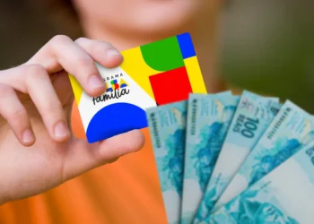 Cartão do programa Bolsa Família sendo segurado por uma pessoa ao lado de notas de dinheiro, representando a antecipação do benefício em cidades brasileiras.