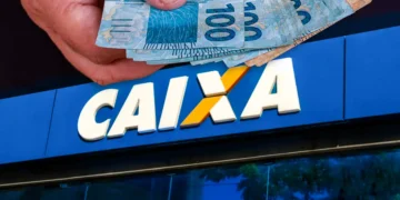 Mão segura cédulas de R$ 100 em frente à fachada da CAIXA