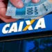 Mão segura cédulas de R$ 100 em frente à fachada da CAIXA