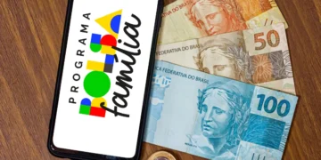 Smartphone exibindo o logotipo do Programa Bolsa Família sobre cédulas de R$ 20, R$ 50 e R$ 100 representando o calendário de pagamento de março de 2026