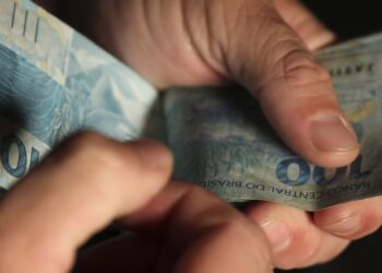 Mãos contando cédulas de R$ 100 reais em fundo escuro — 13º salário do INSS antecipação para aposentados em 2026
