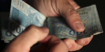 Mãos contando cédulas de R$ 100 reais em fundo escuro — 13º salário do INSS antecipação para aposentados em 2026