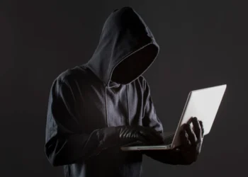 Um hacker com capô e luvas usando um laptop, simbolizando o perigo de golpes online relacionados a taxas para liberação de benefícios.