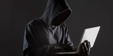 Um hacker com capô e luvas usando um laptop, simbolizando o perigo de golpes online relacionados a taxas para liberação de benefícios.