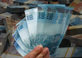 ão segura quatro notas de R$ 100 em frente a pilhas de dinheiro