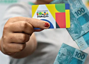 Uma pessoa segurando um cartão do Bolsa Família junto a cédulas de R$ 100, ilustrando o pagamento antecipado do benefício.