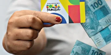 Uma pessoa segurando um cartão do Bolsa Família junto a cédulas de R$ 100, ilustrando o pagamento antecipado do benefício.