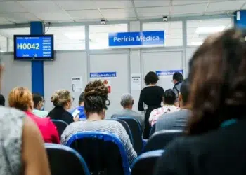 Pessoas aguardando atendimento em uma unidade de perícia médica, com atendentes sendo chamados na tela, representando o mutirão em março.