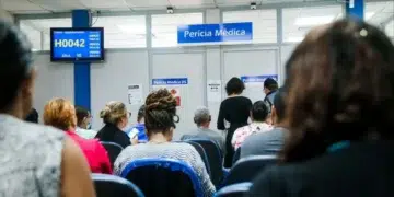 Pessoas aguardando atendimento em uma unidade de perícia médica, com atendentes sendo chamados na tela, representando o mutirão em março.