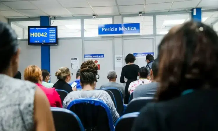 Pessoas aguardando atendimento em uma unidade de perícia médica, com atendentes sendo chamados na tela, representando o mutirão em março.