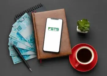 Imagem mostrando cédulas de R$100, um smartphone com o logotipo do FGTS, uma xícara de café e uma planta sobre uma mesa.