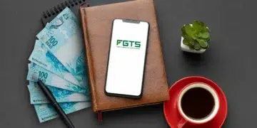 Imagem mostrando cédulas de R$100, um smartphone com o logotipo do FGTS, uma xícara de café e uma planta sobre uma mesa.
