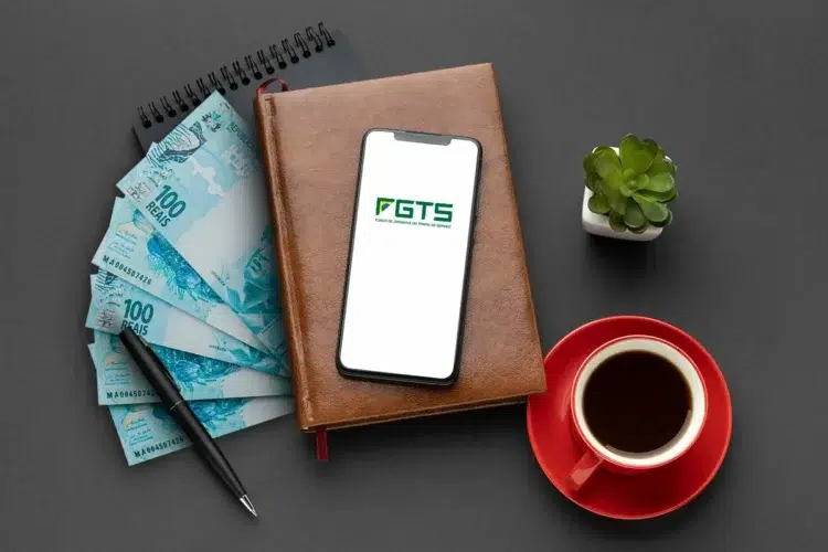 Saque-Aniversário: aniversariantes de março já podem resgatar parcela anual 1 Imagem mostrando cédulas de R$100, um smartphone com o logotipo do FGTS, uma xícara de café e uma planta sobre uma mesa.