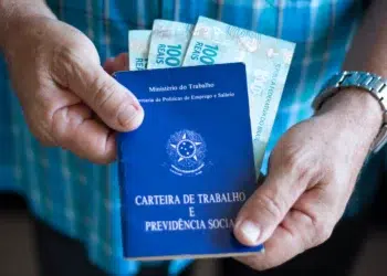 Mãos masculinas segurando carteira de trabalho azul com notas de R$ 100 e relógio prateado no pulso.