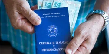 Mãos masculinas segurando carteira de trabalho azul com notas de R$ 100 e relógio prateado no pulso.
