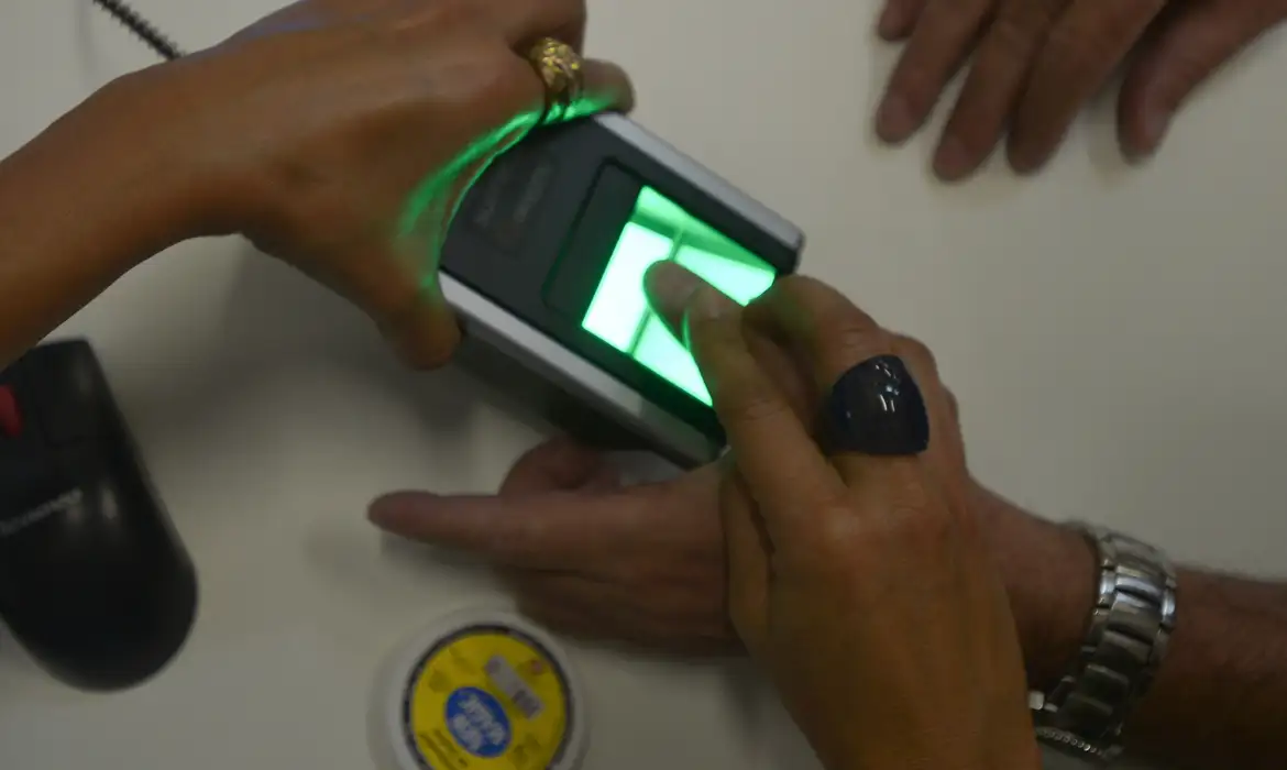 Nova exigência: biometria na identidade será necessária para acesso a programas sociais 2 Mãos realizando coleta de impressão digital em leitor biométrico com luz verde em ambiente de atendimento