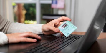 Mãos femininas seguram carteira de identidade verde com foto, em frente a notebook preto sobre mesa de madeira