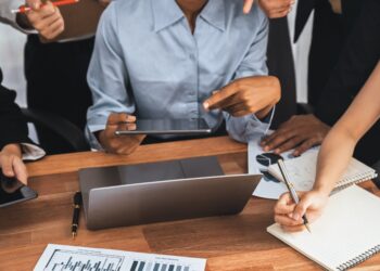 Profissionais em reunião corporativa analisando gráficos e fazendo anotações em mesa de madeira com laptop