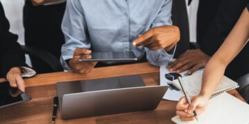 Profissionais em reunião corporativa analisando gráficos e fazendo anotações em mesa de madeira com laptop