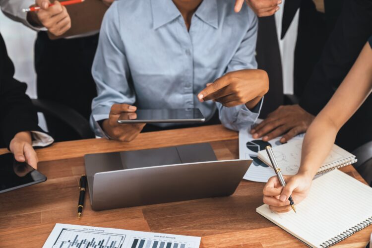 Profissionais em reunião corporativa analisando gráficos e fazendo anotações em mesa de madeira com laptop