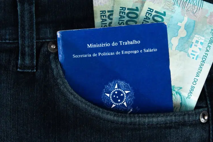 Novo grupo é incluído no auxílio-creche de R$ 526,64; confira se você tem direito 2 Carteira de trabalho azul do Ministério do Trabalho em bolso jeans escuro junto com notas de R$ 100.