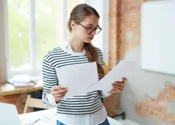 Jovem de óculos com camisa listrada segurando papéis em escritório iluminado