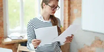 Jovem de óculos com camisa listrada segurando papéis em escritório iluminado