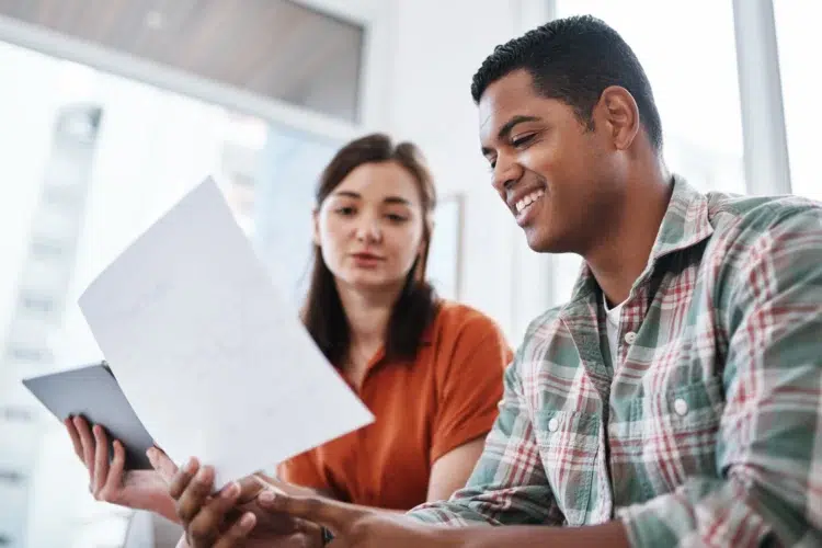 Governo reajusta bolsa-estágio em 33% e alcança 14.800 estudantes 2 Dois jovens sentados analisando juntos uma folha de papel e um tablet em escritório iluminado