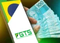 Mão segurando celular com aplicativo FGTS e bandeira do Brasil ao lado de cédulas de R$ 100.