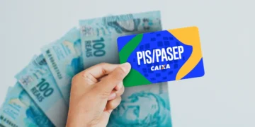 Mão segurando cartão PIS/Pasep Caixa acima de várias notas de 100 reais