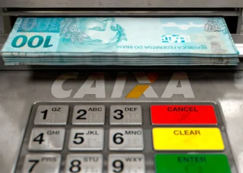 Notas de R$ 100 saindo de caixa eletrônico da Caixa com teclado numérico e botões coloridos visíveis.