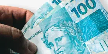 Mão segura cédulas de R$ 100 reais com efígie da República, simbolizando alta de 5,9% no salário mínimo prevista para 2027.