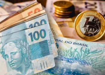 Notas de R$ 100 e R$ 50 empilhadas ao lado de moedas de R$ 1 sobre fundo com cédulas de real brasileiras.