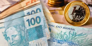 Notas de R$ 100 e R$ 50 empilhadas ao lado de moedas de R$ 1 sobre fundo com cédulas de real brasileiras.