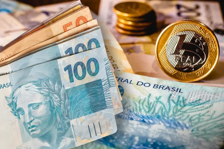 Notas de R$ 100 e R$ 50 empilhadas ao lado de moedas de R$ 1 sobre fundo com cédulas de real brasileiras.
